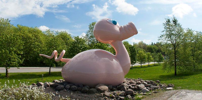 A huge pink pig-duck art piece on top of small rocks, surrounded by green trees and grass
