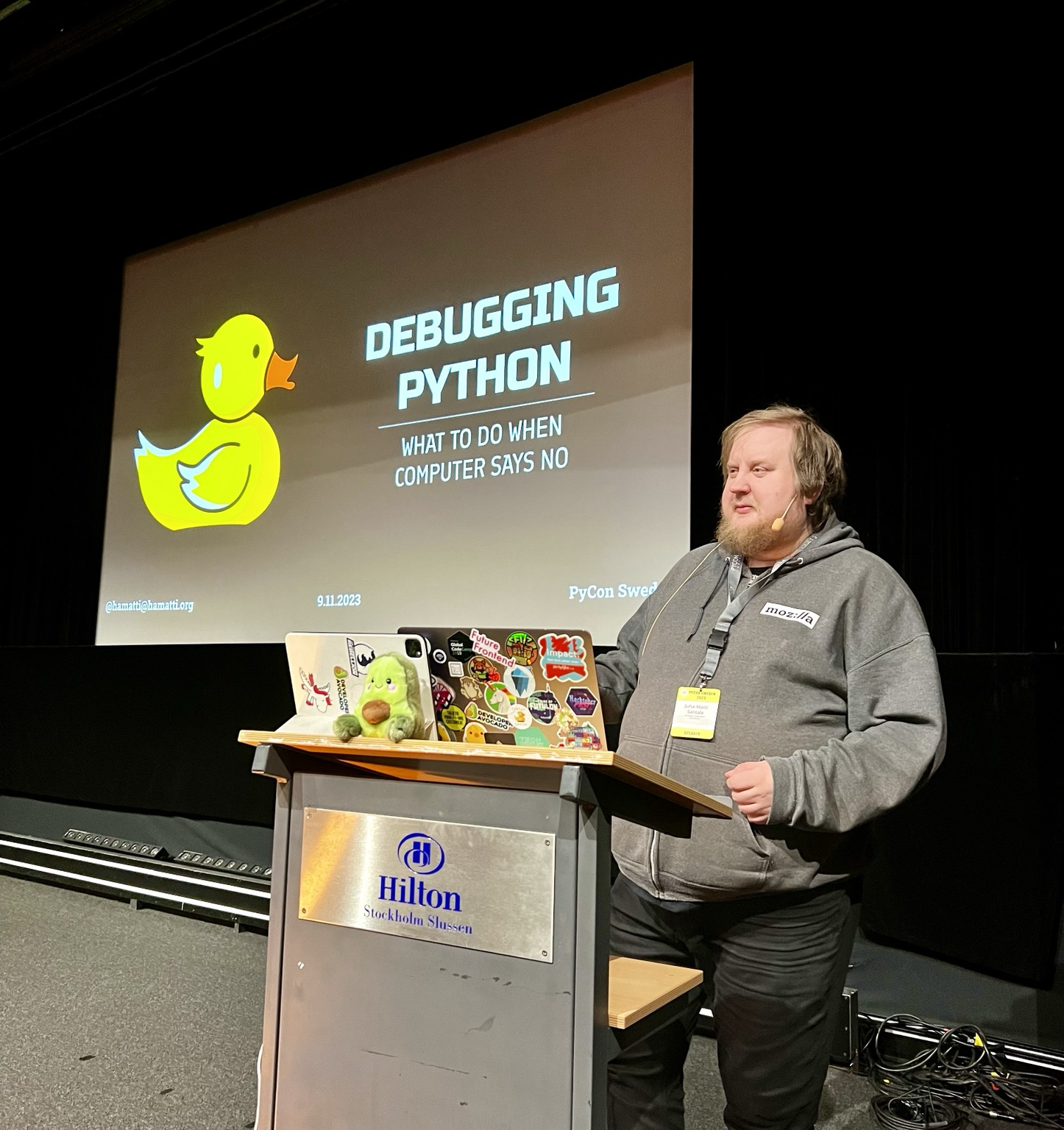 Juhis on stage standing behind a speaker podium. On the podium, there's a laptop, an iPad and a cute avocado plushie. Juhis is wearing a grey hoodie and black jeans. On the background, a projector screen with a slide that has a yellow rubber duck illustration and title "Debugging Python - what to do when computer says no".
